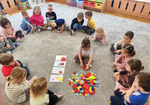 Dzieci siedzą w kole na dywanie. Na środku dywanu rozłożone są karty edukacyjne, plastikowe figury. Dziecko wyszukuje wybrane elementy.