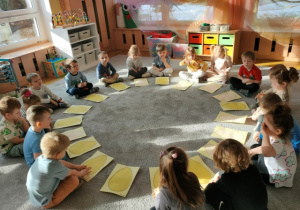 Dzieci siedzą w dużym kole na dywanie, na którym przed nimi rozłożone są ich prace.