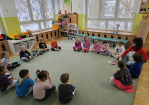 Dzieci siedzą na dywanie i zaciekawienie słuchają opowiadania czytanego przez Panią Anię.