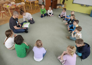 Dzieci siedzą na dywanie a tata Małgosi czyta opowiadanie.