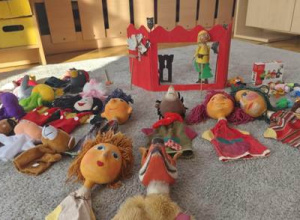 Przed mini teatrzykiem stojącym na dywanie rozłożone są różne kukiełki i pacynki.
