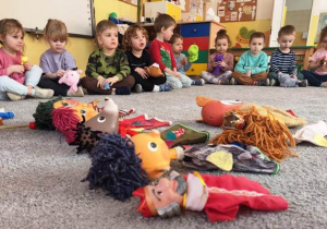 Dzieci oglądają pacynki i kukiełki zgromadzone na dywanie.