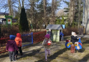 Dzieci bawią się - dziewczynka biegnie z piłka, inna buja się na bujaku, chłopcy idą do domku ogrodowego.