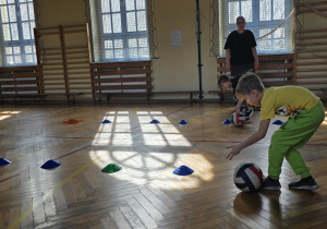 Dzieci turlają piłkę miedzy pachołkami.