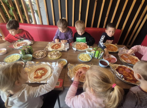 Dzieci siedzą przy stołach i tworzą swoją pizzę.