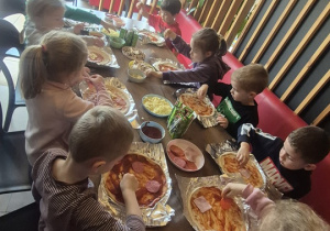 Dzieci siedzą przy stołach i nakładają na swoje Pizze dodatki..