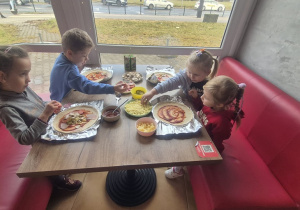 Dzieci nakładają na pizzę dodatki takie jak ogórki, pieczarki.