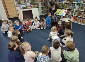 Dzieci siedzą w czytelni w kole na dywanie i słuchają czytanej bajki.