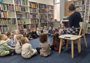 Dzieci siedzą na dywanie w czytelni, słuchają bajki czytanej przez panią bibliotekarkę.