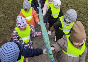 Dzieci idą po chodniku w parach trzymając się węża spacerowego.