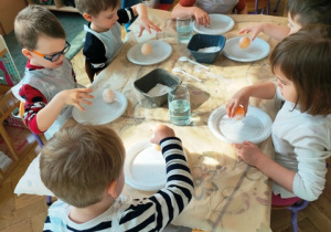 Dzieci siedzą przy stolikach. Sprawdzają czy jajko z odwapnioną skorupką odbija się jak piłeczka.