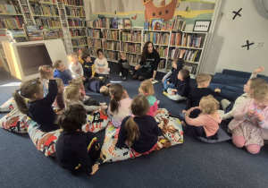 Dzieci siedzą na dywanie w bibliotece i słuchają czytanej bajki o żabach.
