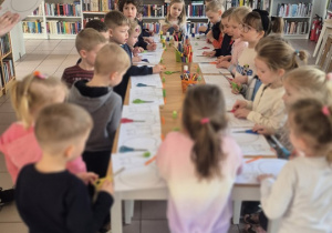 Dzieci stoją przy stołach i wykonują swoje prace plastyczne.