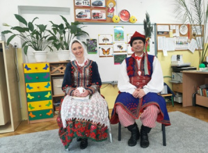 Goście ubrani w krakowskich strojach ludowych siedzą na krzesłach przed dziećmi.