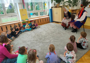 Dzieci siedzą w półkolu na dywanie i słuchają wypowiedzi przybyłych gości.