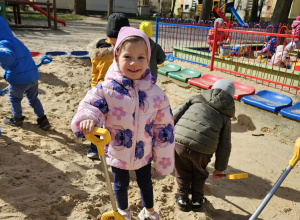 Dziewczynka stoi uśmiechnięta w piaskownicy.