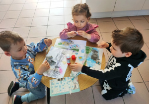 Troje dzieci przykleja do kartki kawałki gazet- budują tamę na rzece.