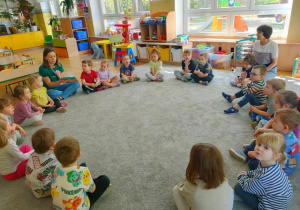 Dzieci siedzą na dywanie w dużym kole. Słuchają wypowiedzi gościa.