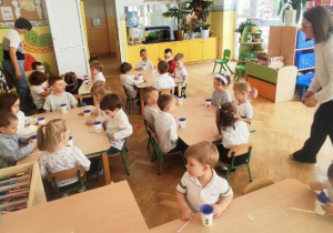 Dzieci siedzą przy stoliczkach, na których naszykowane są: papierowe kubeczki z dziurką z nałożonymi na nie rękawiczkami lateksowymi, oraz słomki .