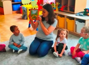 Mama kolegi siedzi na dywanie przed dziećmi i prezentuje dzieciom książkę o Franklinie.