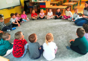 Dzieci siedzą na dywanie. Mama kolegi prezentuje dzieciom książkę, którą będzie czytać.