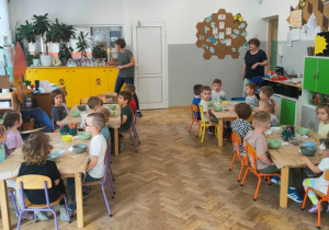 Dzieci siedzą przy stolikach i słuchają prowadzącej, która wyjaśnia dzieciom ich zadanie.