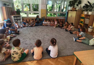 Mama siedzi na dywanie wraz z dziećmi w dużym kole. Prezentuje przez siebie przyniesioną książkę.
