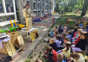 Dzieci siedzą na piankach w ogrodzie czekając na rozpoczęcie spotkania.