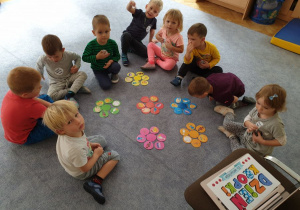 Dzieci siedzą w kole na dywanie i grupują kolorowe krążki zgodnie z ich barwą.