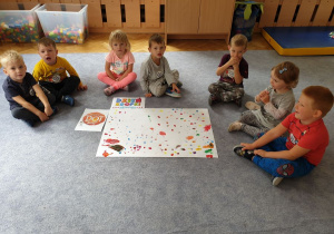 Dzieci siedzą na dywanie w półkolu, a przed nimi leży ich praca plastyczna z kolorowymi kropkami.