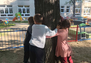 Troje dzieci przytulają się do drzewa w ogrodzie.