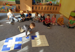 Chłopiec układa na kratownicy kolorowe kwadraty zgodnie z ułożonym koło niego wzorem.