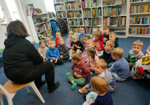 Dzieci siedzą na dywanie i rozmawiają z panią bibliotekarką. Odpowiadają na jej pytania.