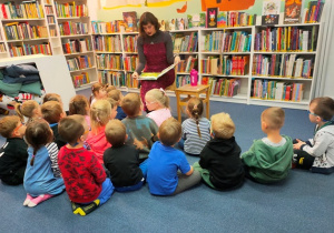 Dzieci siedzą na dywanie w czytelni i słuchają bajki czytanej przez panią bibliotekarkę.