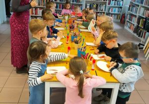 Dzieci wykonują prace plastyczną stojąc przy stolikach na krórych rozłożone są wszystkie potrzebne materiały plastyczne i przybory.
