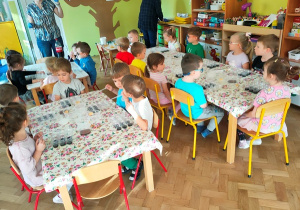 Dzieci siedzą przy stolikach na których ustawione są różne pojemniki z cieczami.