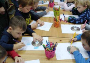 Dzieci stoją przy stole i rysują po kartkach papieru.