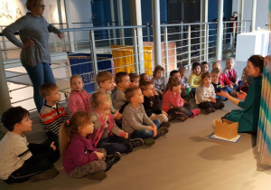Dzieci siedząc na podłodze słuchają wypowiedzi przewodnika o różnych kolorach ukrytych w tkaninach.