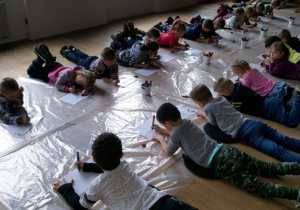 Dzieci leżąc na brzuchu tworzą prace plastyczną na arkuszach przezroczystej folii.