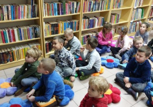 Dzieci siedzą na poduszkach na podłodze wśród książek w bibliotece.