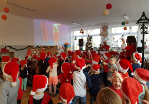 Dzieci ubrane w czerwone czapeczki Mikołaja tańczą do wesołej melodii.