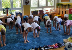 Dzieci wykonują skłon w przód, rękami próbują dotknąć dywanu.