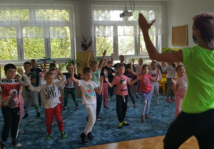 Dzieci stoją w luźnej grupie na dywanie. Z uniesionymi do góry rękami w rytm muzyki poruszają nogą.