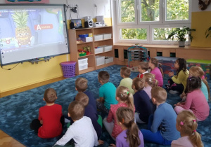 Dzieci siedzą w siadzie skrzyżnym na dywanie i oglądają bajkę o literce A wyświetlaną na tablicy multimedialnej.