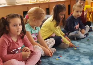 Dzieci siedzą w siadzie skrzyżnym na dywanie. Przed nimi leżą krążki odpowiadające liczbie sylab w podanym przez nauczycielkę słowie.