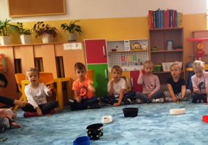 Dzieci siedzą w dużym kole na dywanie. Dzielą słowa na sylaby pokazując ilość sylab na palcach.