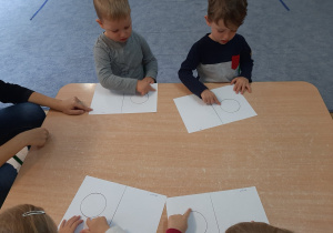 Dzieci siedzą przy stoliku i rysują paluszkiem po śladzie.