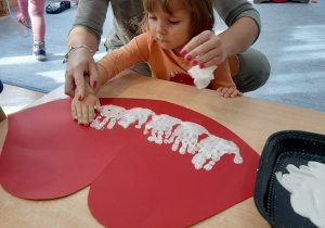 Dziewczynka odbija rączkę pomalowaną białą farbą na ustach wyciętych z papieru tworząc zęby.