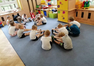 Dzieci siedzą na dywanie i wykonują skłon.