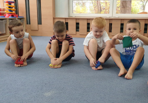 Chłopcy siedzą na dywanie i próbują unosić stopy wraz z woreczkiem do góry.